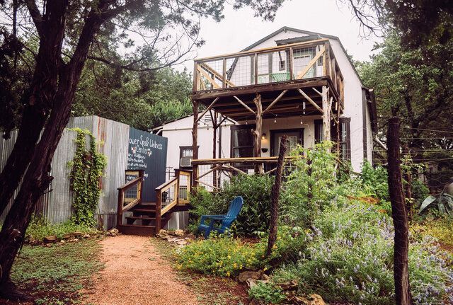 A small white house with a wooden deck is surrounded by trees and bushes.