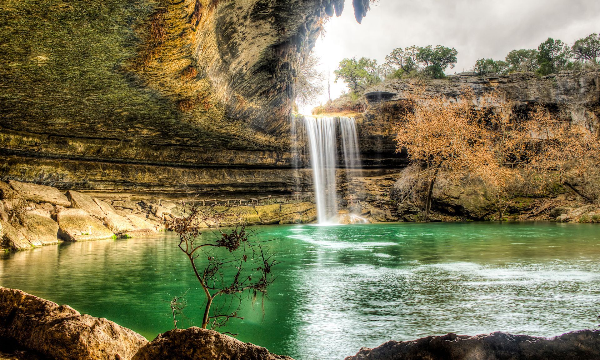 Hamilton Pool Preserve features a waterfall cascading into a vibrant turquoise pool surrounded by limestone cliffs.