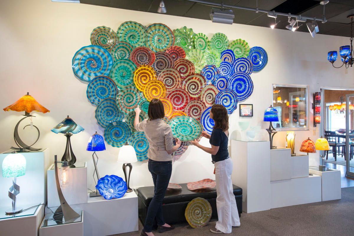 Two people examine a vibrant wall installation of multicolored, spiraled glass plates inside a bright art gallery.