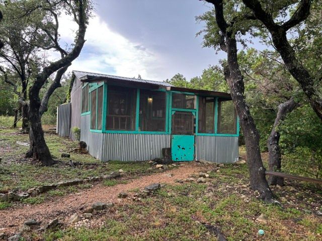 Small turquoise cabin with screened porch in a wooded clearing, surrounded by trees and a dirt path.