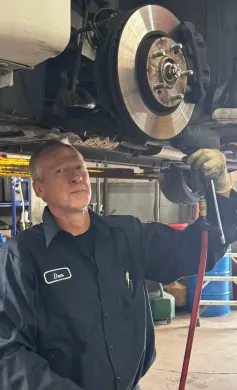 Mechanic working on a car's brakes. Man in blue work shirt, holding tool, under car. Workshop setting. | Loyola Service