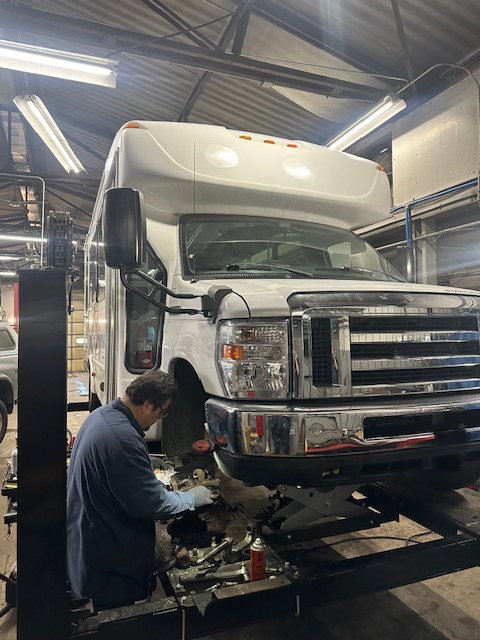 Mechanic working on the front wheel of a white RV raised on a lift inside a garage. | Loyola Service