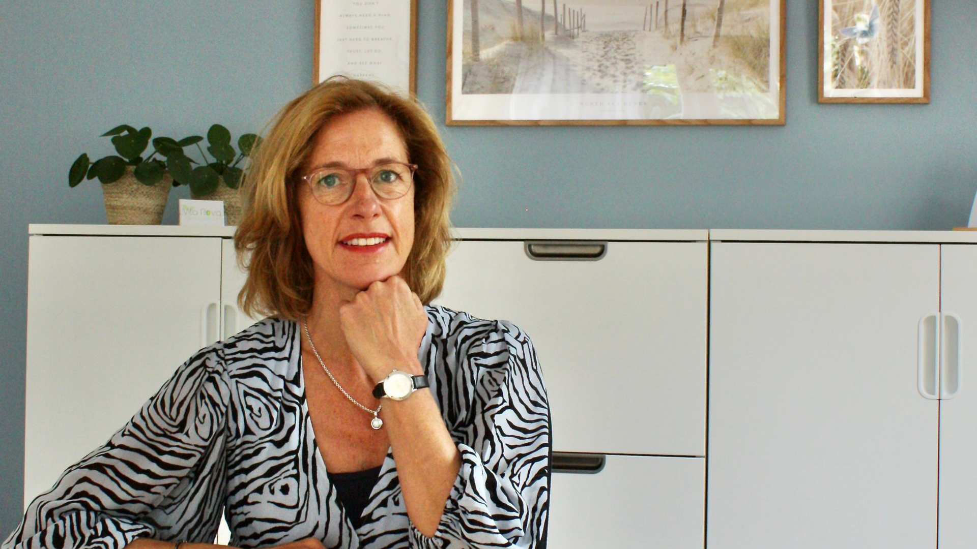 Lachende vrouw met bril zittend aan bureau. Witte kasten op achtergrond