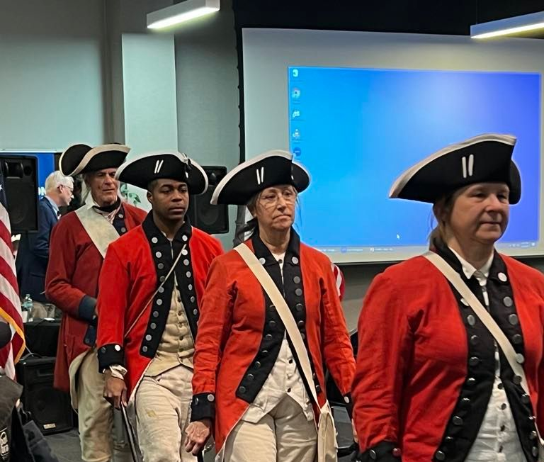 Line of people in Revolutionary War era red coats and hats walking.