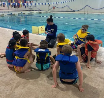 Children in life jackets sit by an indoor pool while an instructor leads a swimming lesson