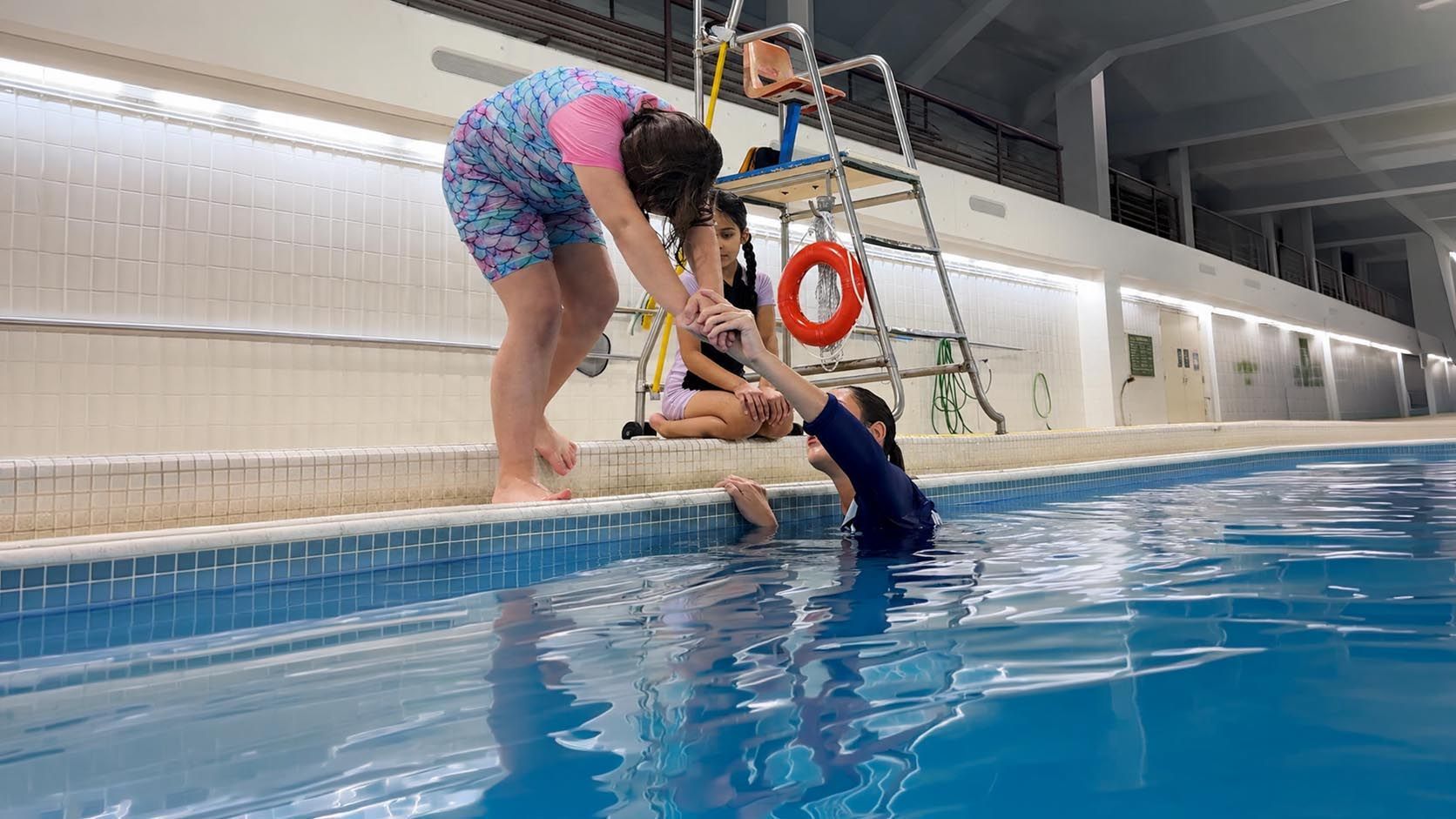 Indoor pool rescue drill with swimmers and a red rescue float being tossed to a person in the water
