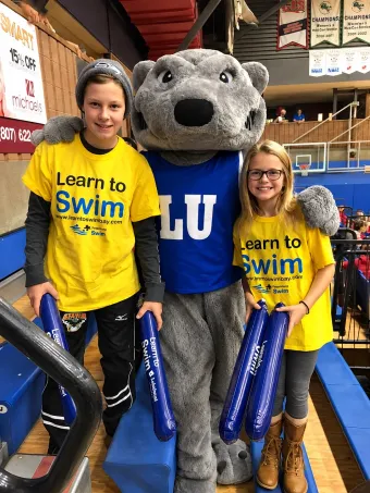 Three people pose with a LU mascot in a gym, wearing yellow and blue Learn to Swim shirts.