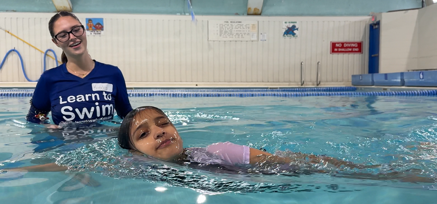 Swimmer and instructor in an indoor pool, with the swimmer floating near the instructor wearing a “Learn to Swim” shirt