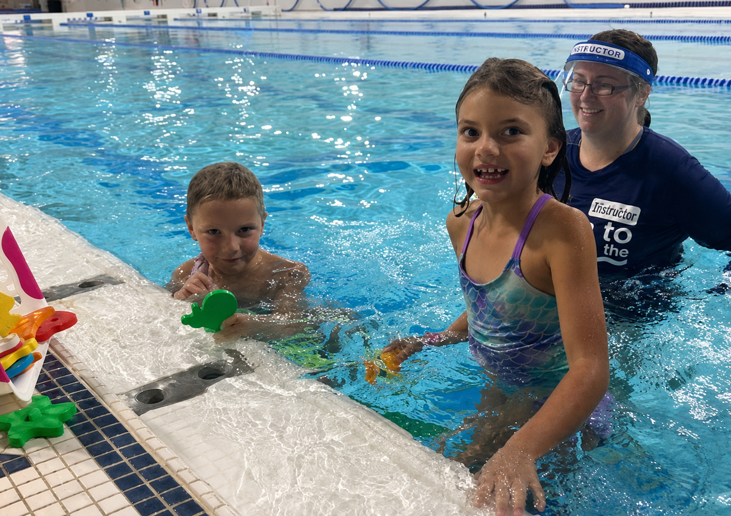Children smiling in a swimming pool with a coach beside them near the pool edge