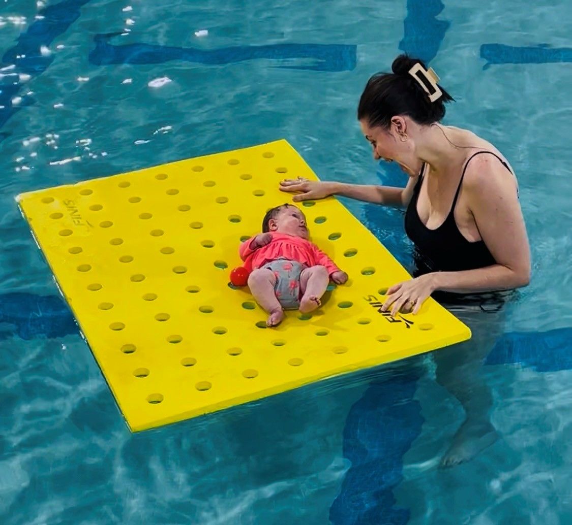 Woman in a pool guiding a baby on a yellow floating mat