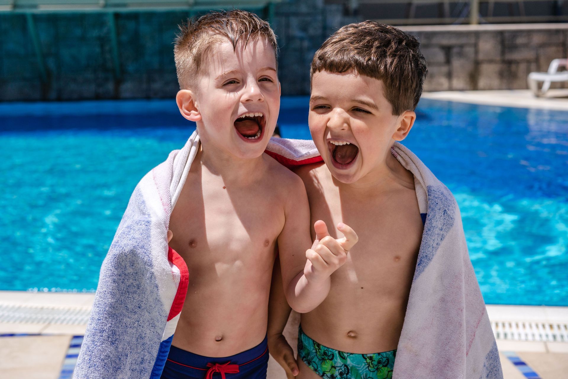 Child smiling on a pool float, reaching toward an instructor in blue in an indoor swimming pool.