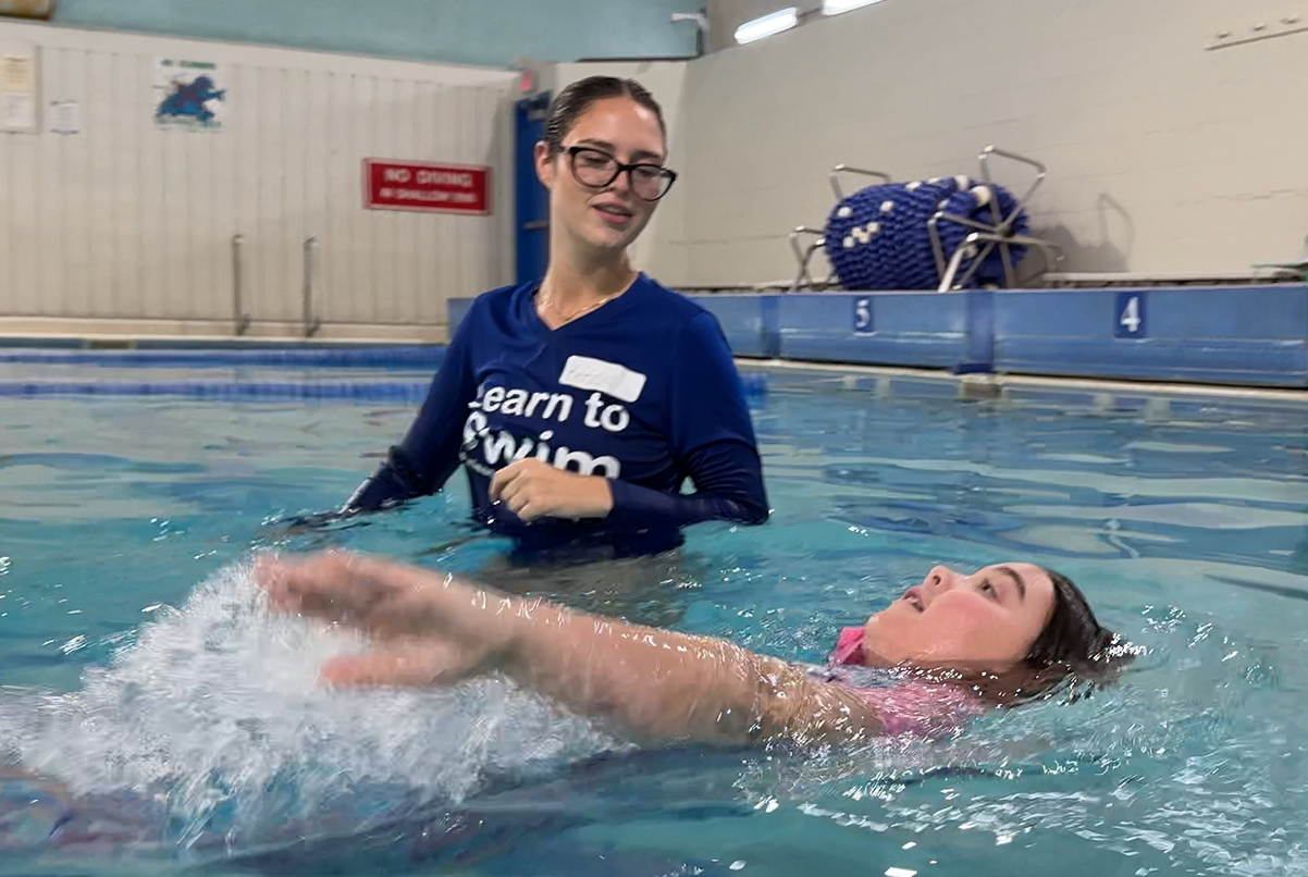 Swim instructor in blue rash guard assists a swimmer floating on their back in an indoor pool