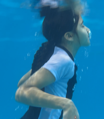 Person swimming underwater in a blue pool, wearing a white shirt.