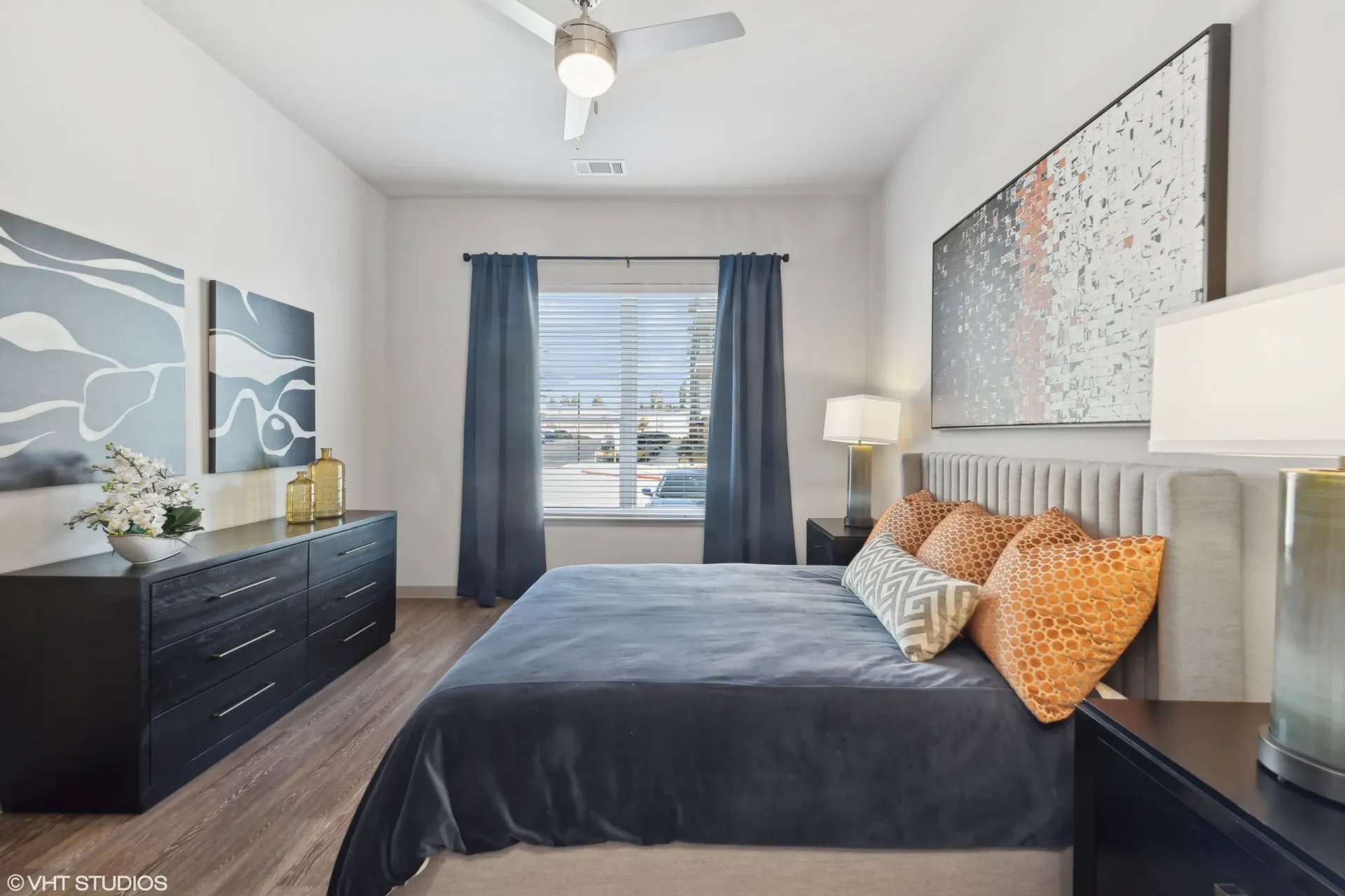 Bedroom with dark bedspread, upholstered headboard, dresser, and a window with blue curtains.
