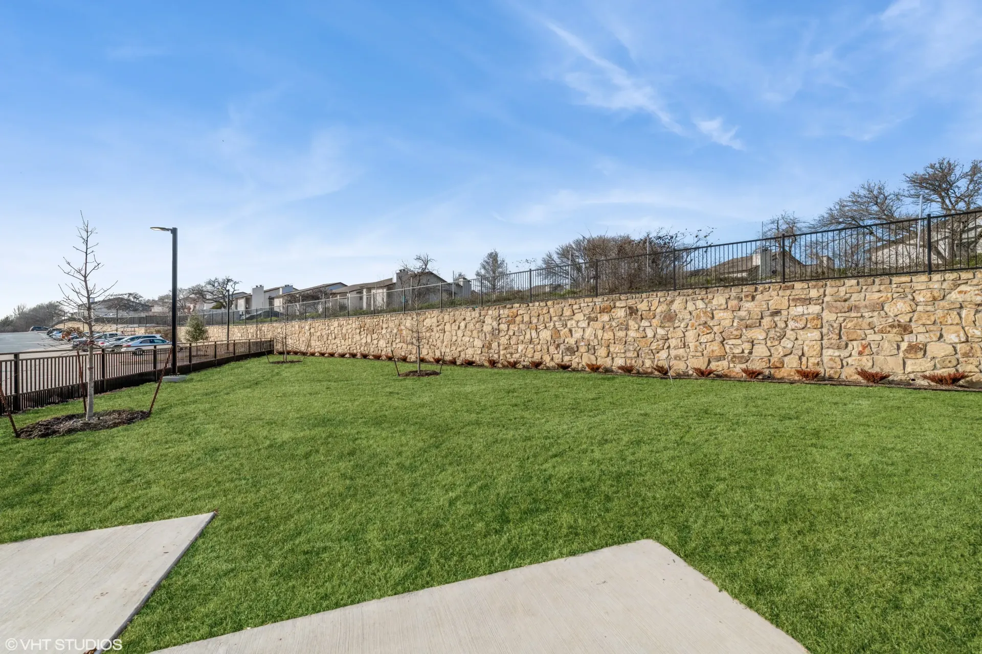 Green lawn with a stone retaining wall, black railing, and adjacent parking area.