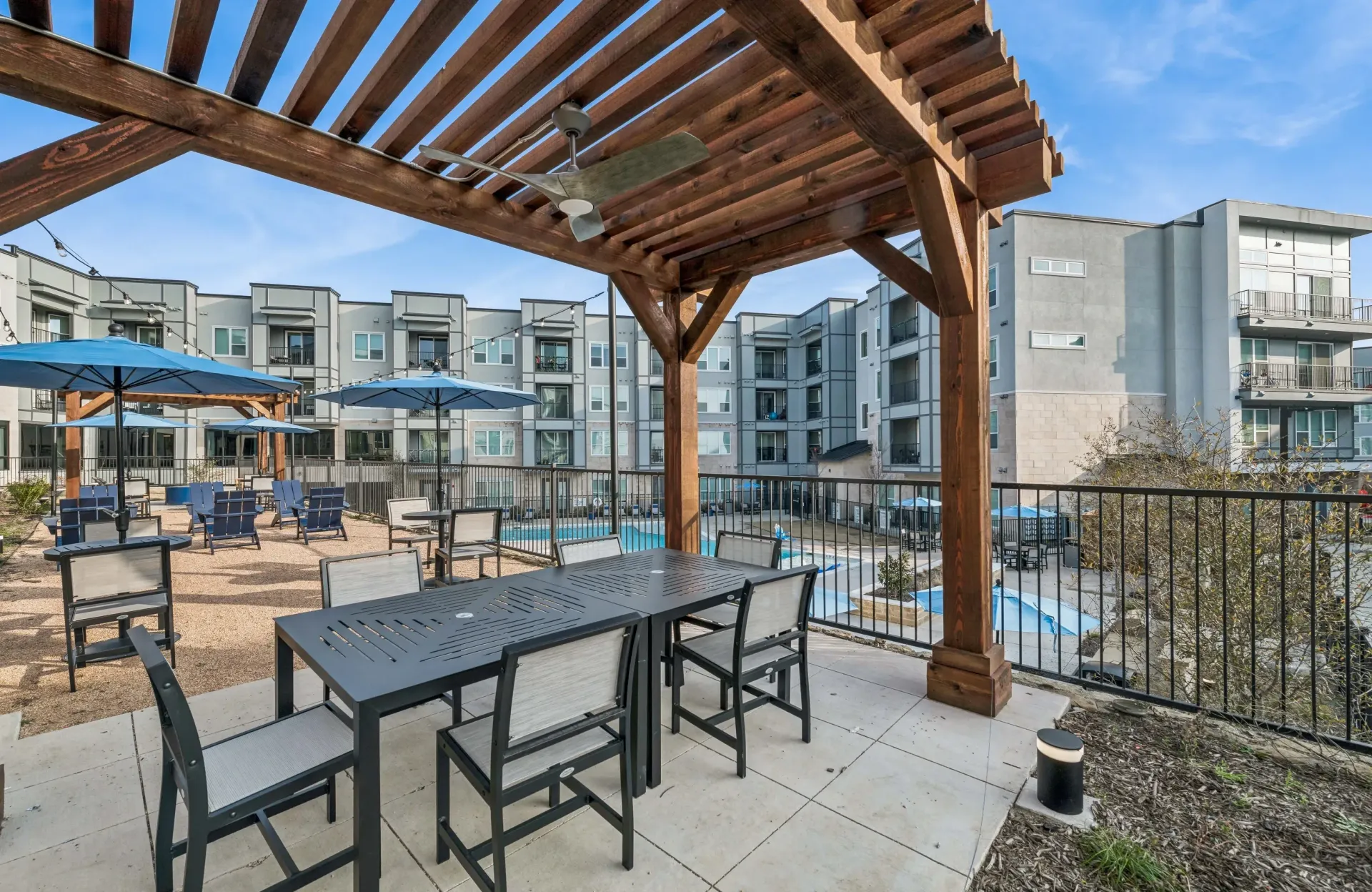 Outdoor community lounge under a wooden pergola with tables and chairs; pool visible in the background.