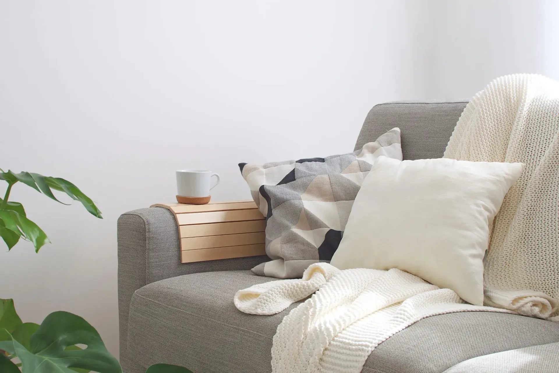 Gray sofa with geometric pillows, a cream knit blanket, a wooden tray with a cup, and a green plant.