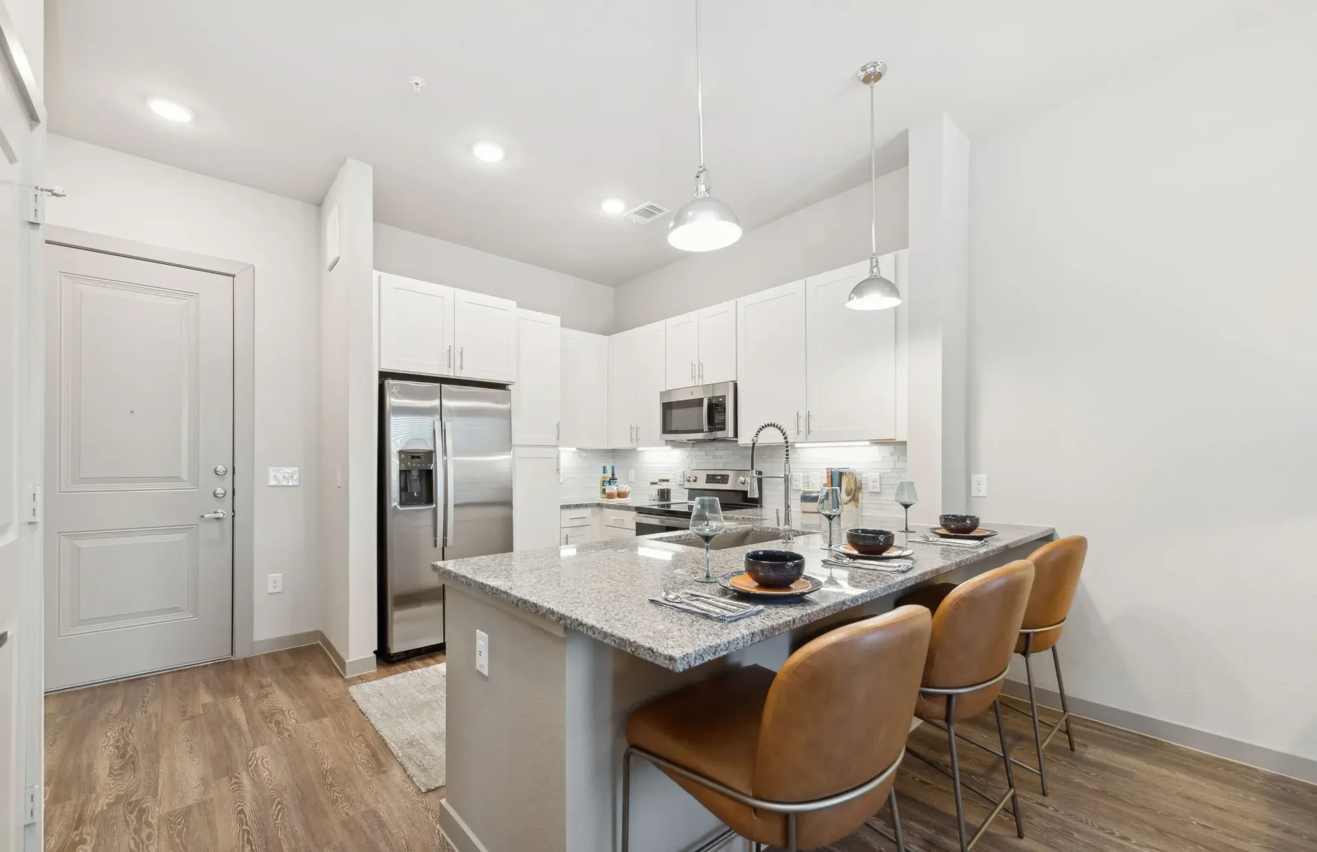 Modern white kitchen with granite island, stainless steel appliances, and four brown leather bar stools.