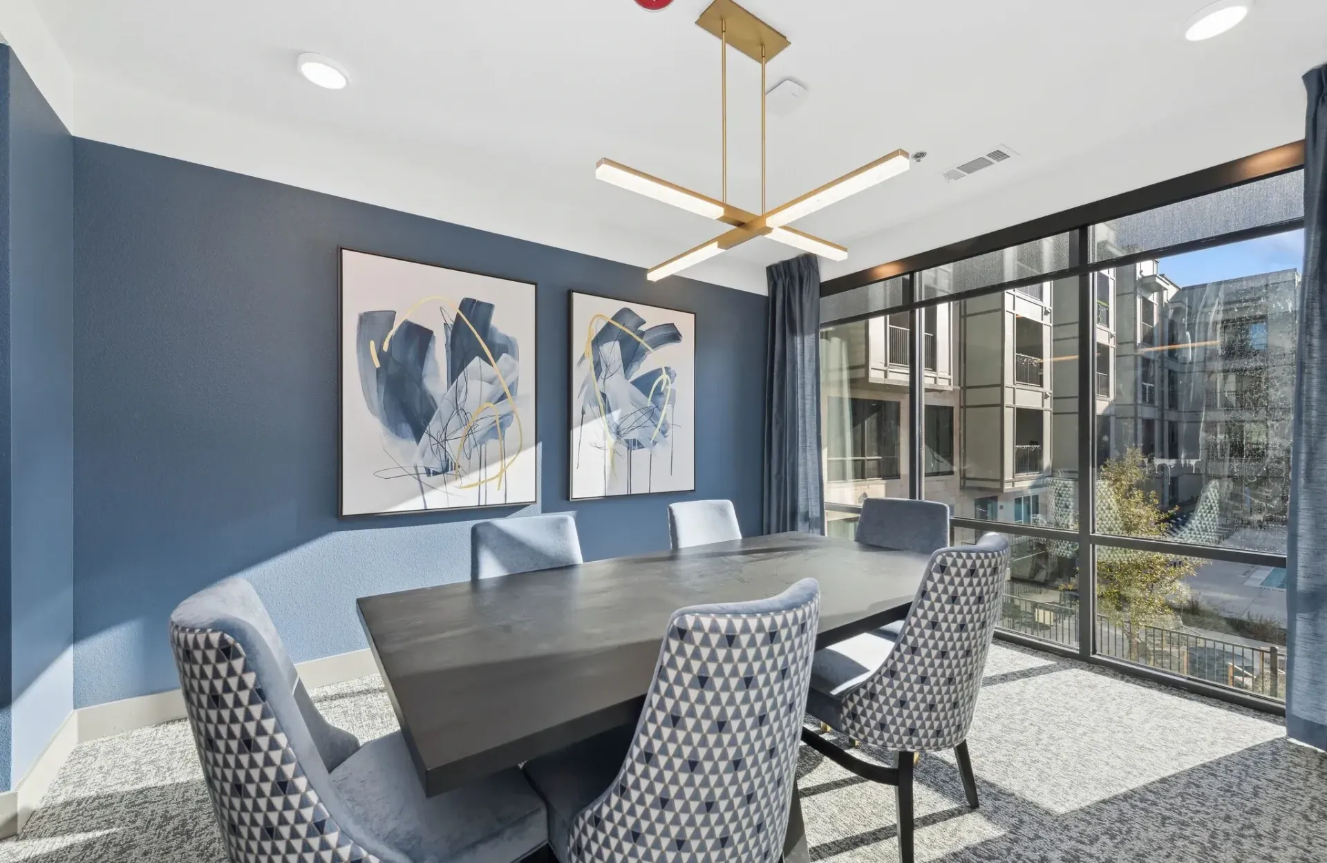 Conference-style community lounge with a dark table, patterned chairs, blue walls, and large windows.