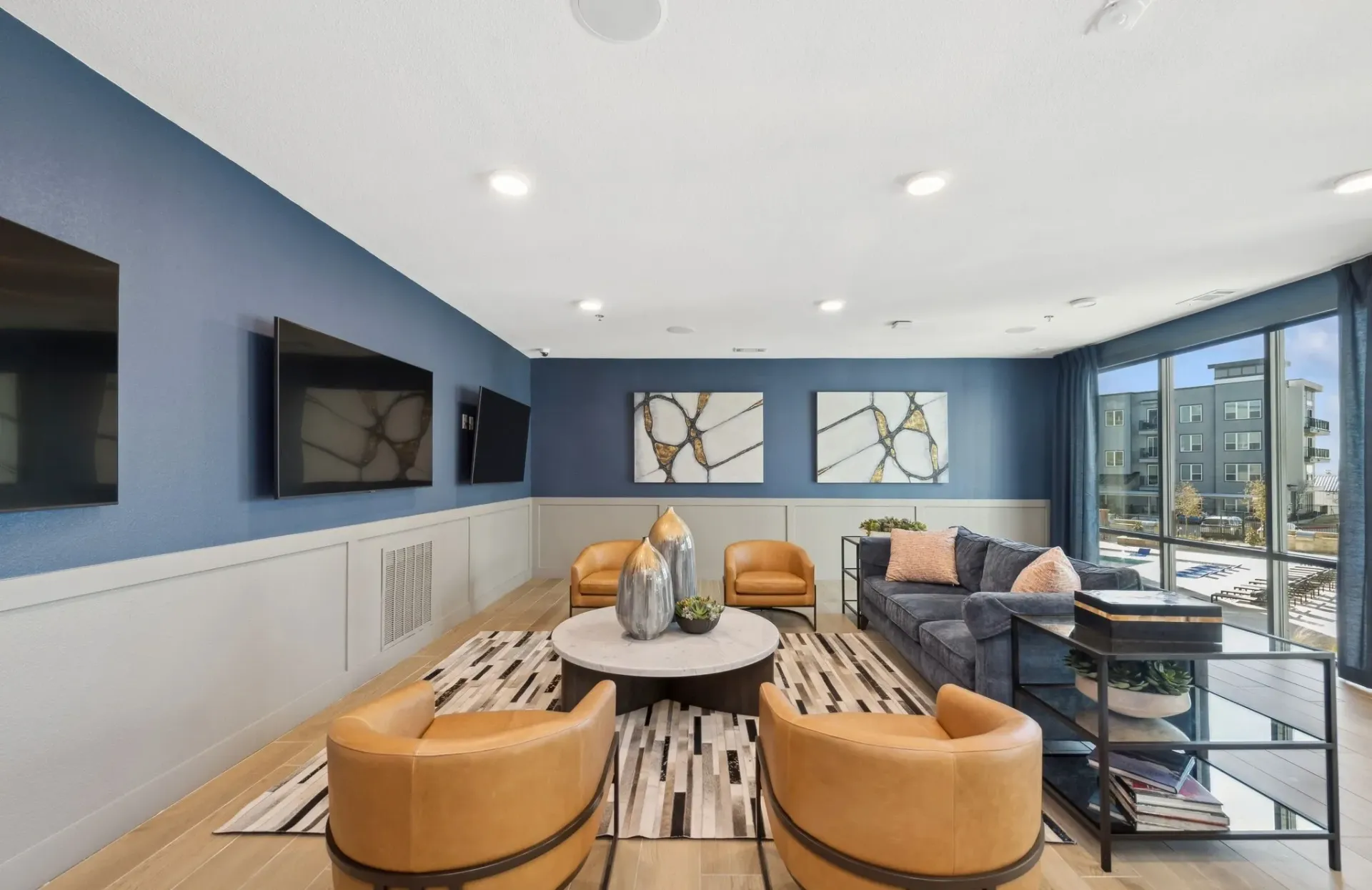 Communal lounge with blue walls, tan chairs, gray sofa, round coffee table, and wall-mounted TVs.