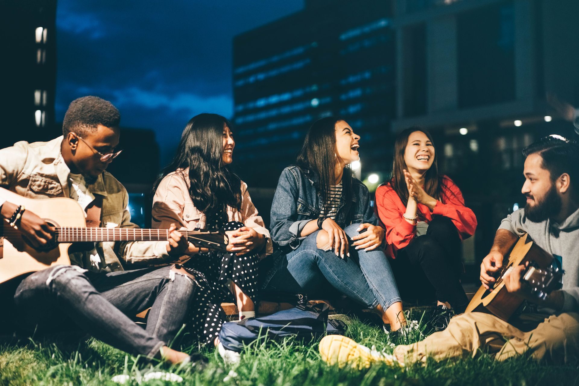 Friends sitting on grass at night, laughing and chatting while playing guitars by city lights