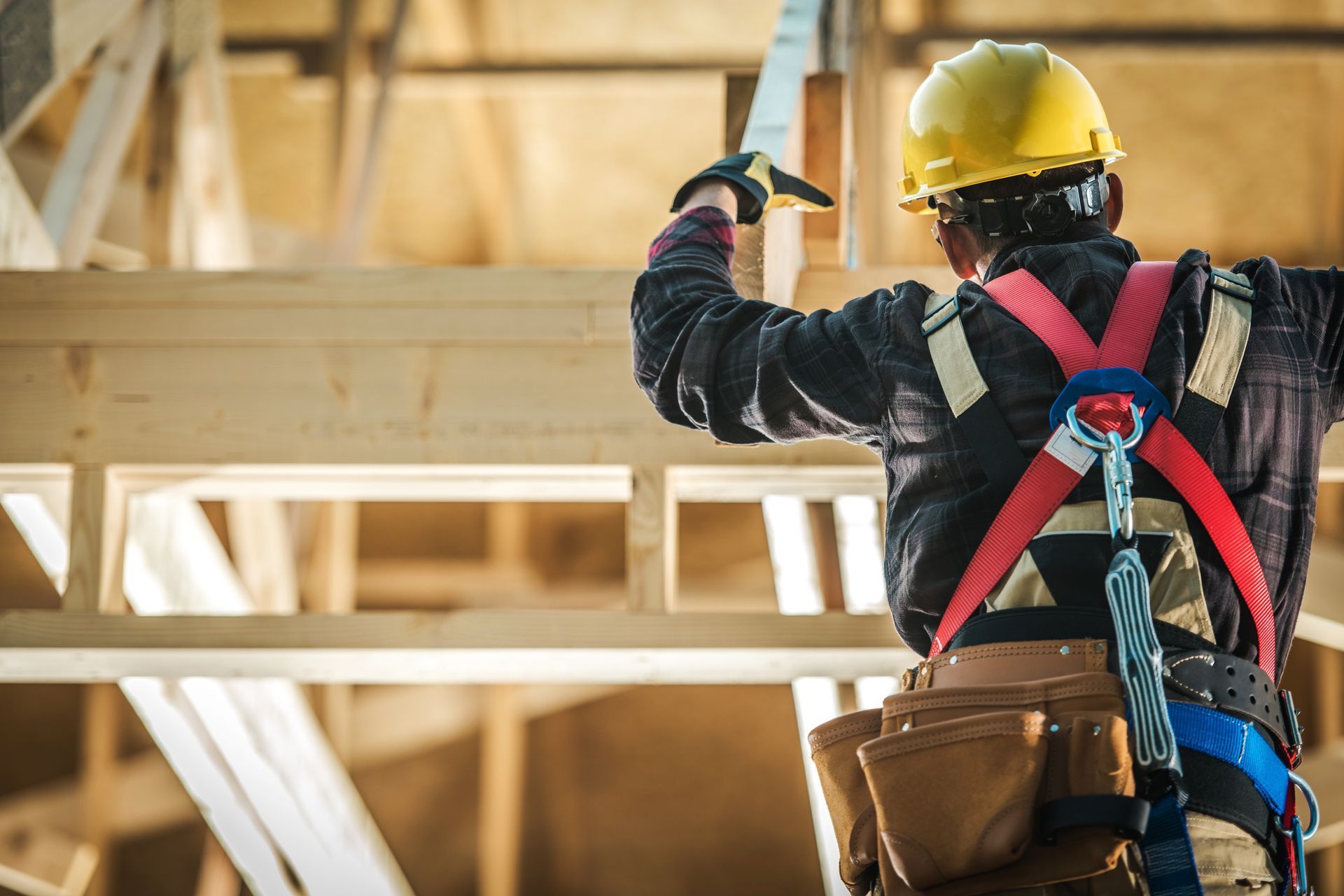 Ouvrier du bâtiment portant un casque jaune, un harnais de sécurité et une ceinture porte-outils, travaillant sur une charpente en bois.