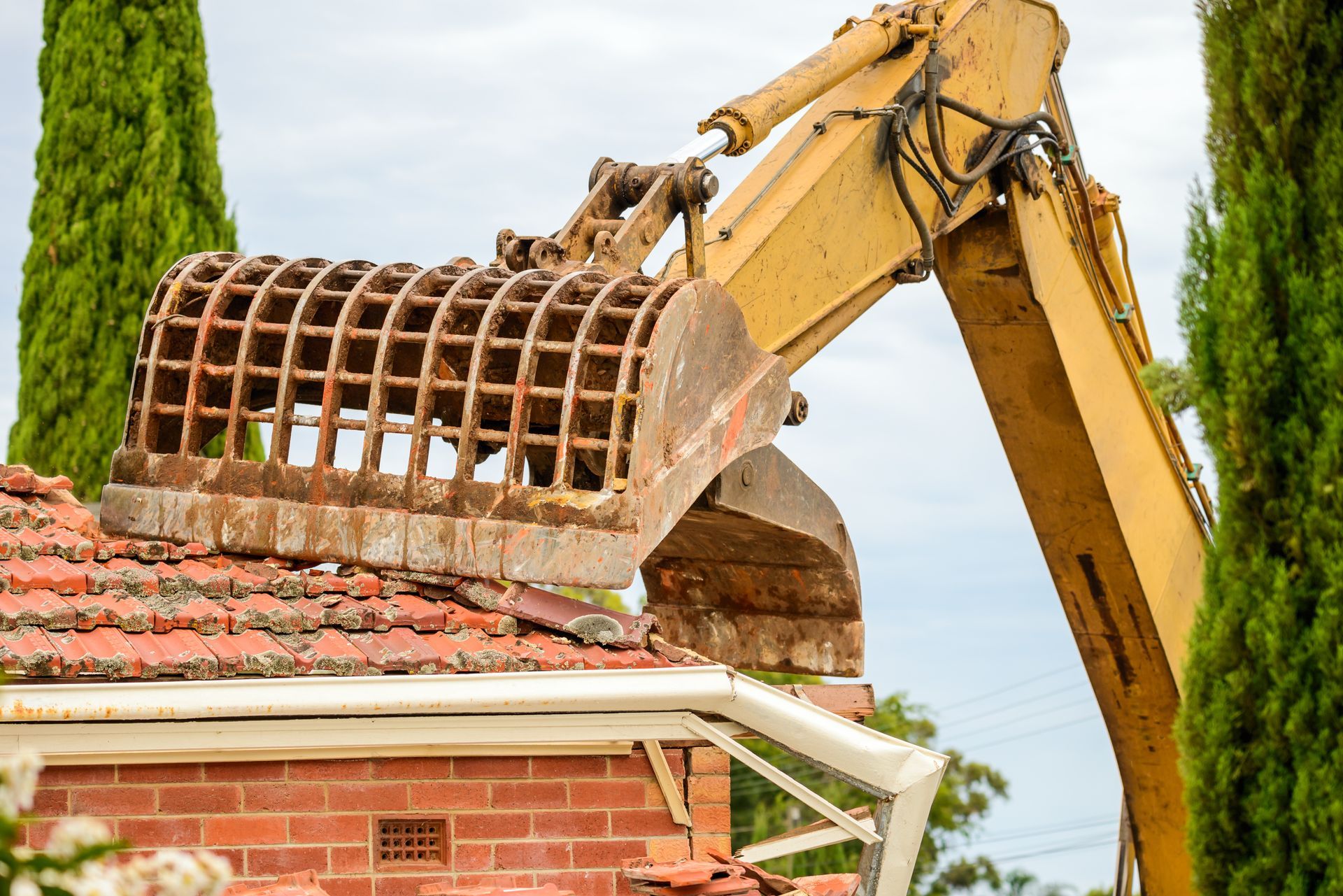 Excavator demolishing a brick house roof beside tall green trees