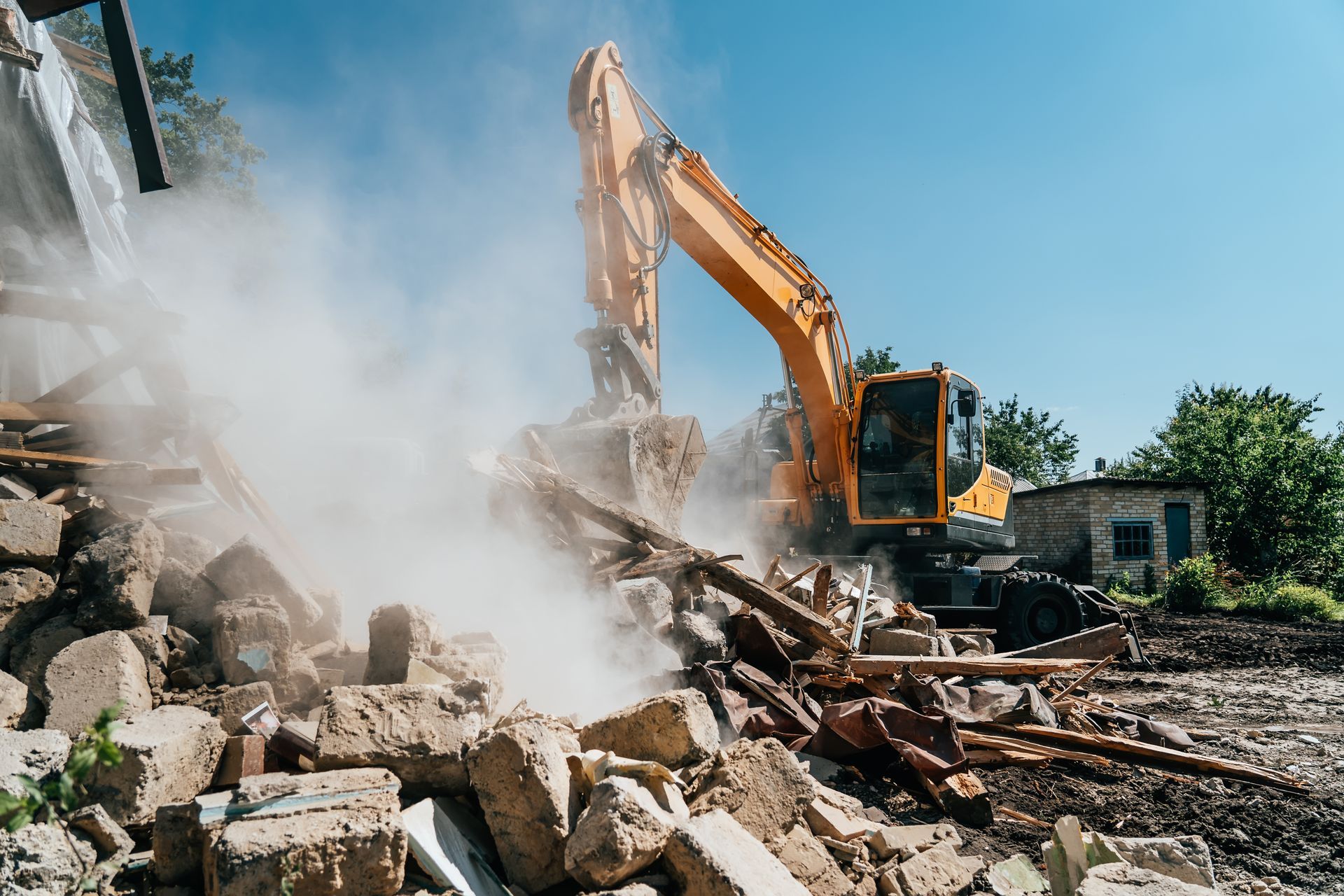Excavator demolishing rubble beside a dusty building, with debris and dust clouds rising.