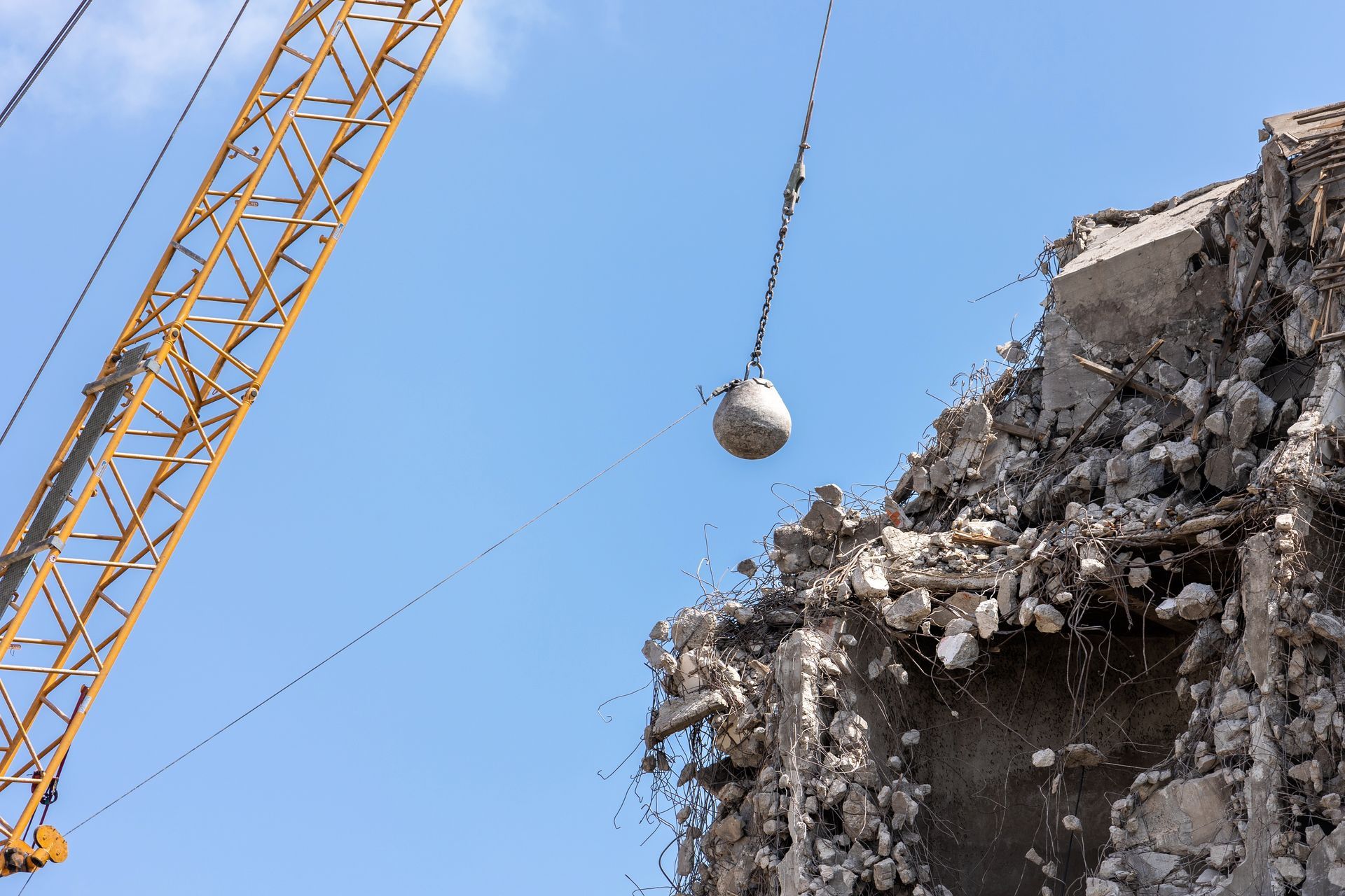 Yellow crane with wrecking ball beside a partially demolished concrete building against blue sky