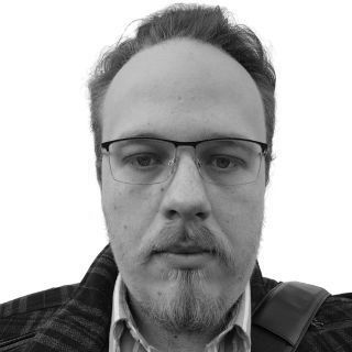 Black-and-white close-up portrait of a man wearing glasses and a jacket, looking at the camera.
