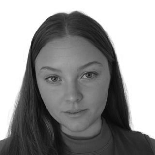 Black-and-white portrait of a person with long straight hair, looking at the camera against a paneled wall.