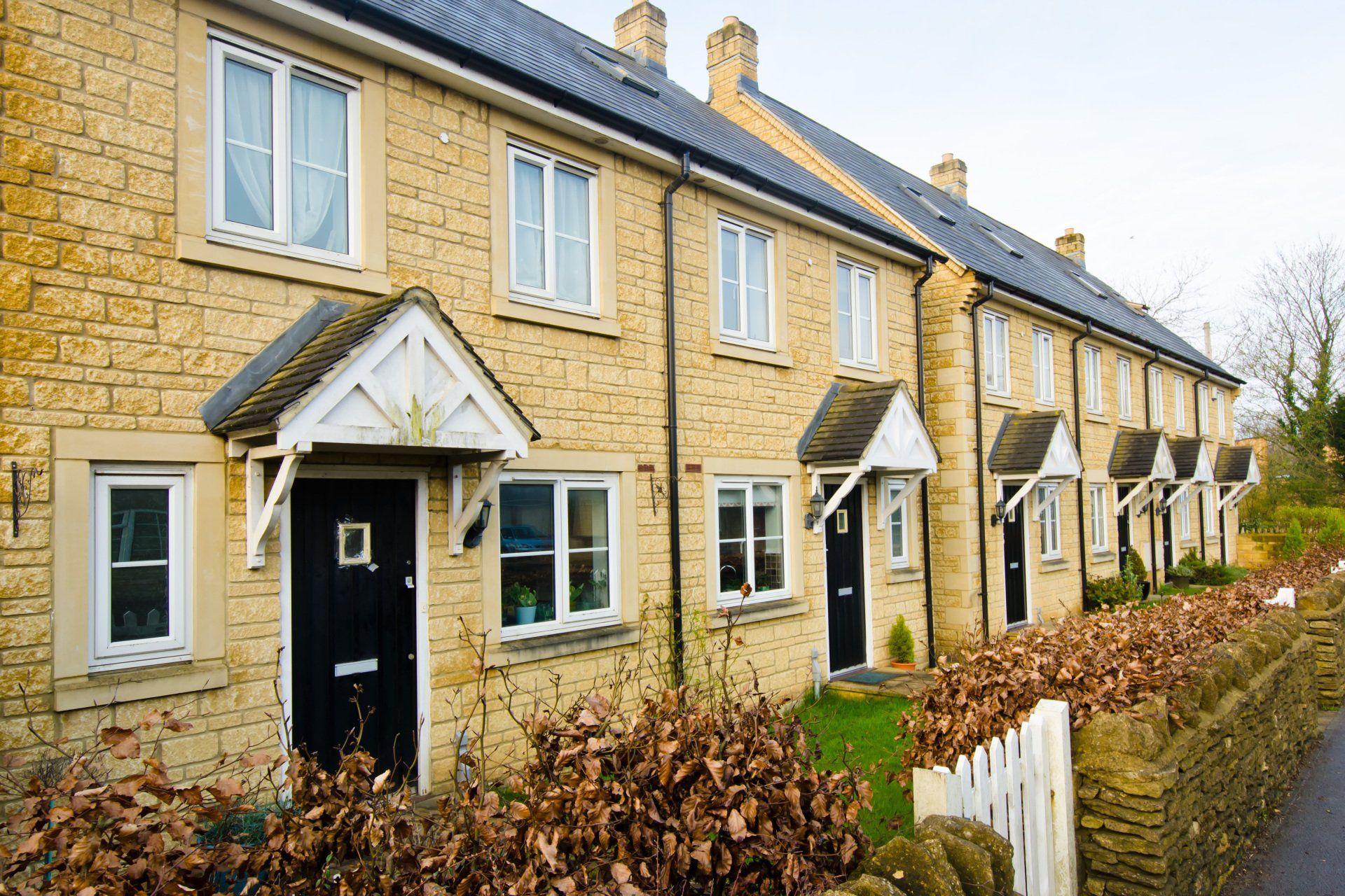 Row of tan stone cottages with black doors and white trim along a leafy street