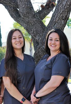 Two women in scrubs are posing for a picture in front of a tree.