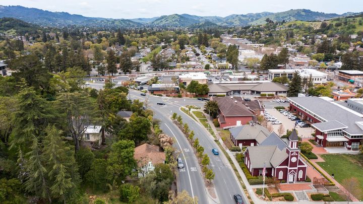 Aerial view of a small town street with buildings, trees, and hills in the distance.