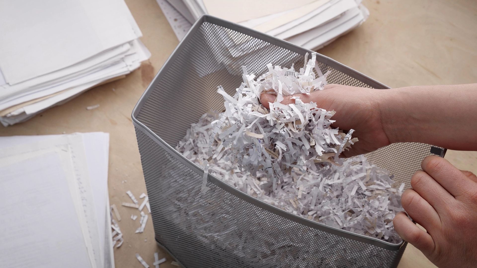 Hands shredding papers into a mesh wastebasket beside stacks of documents