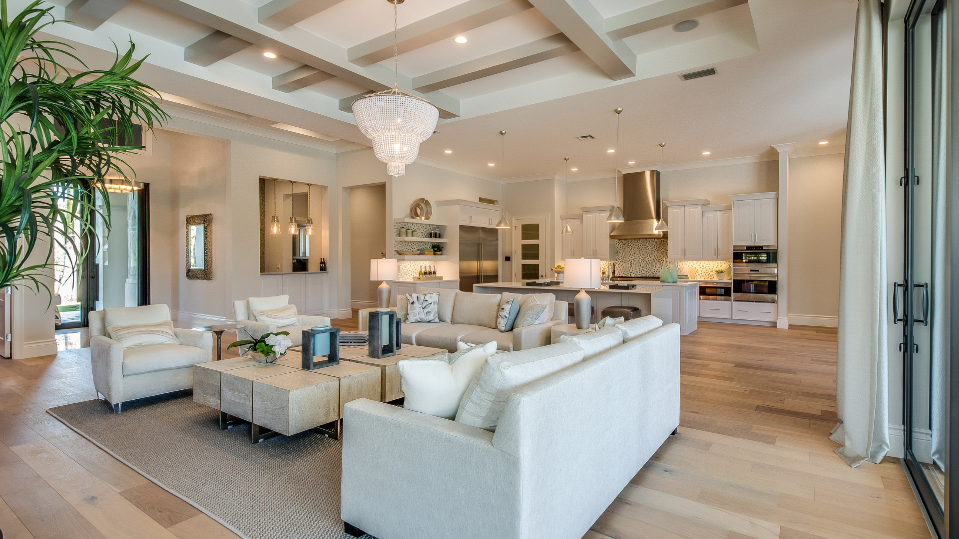 Bright open-concept living room with white sofas, wood beams, chandelier, and kitchen in the background