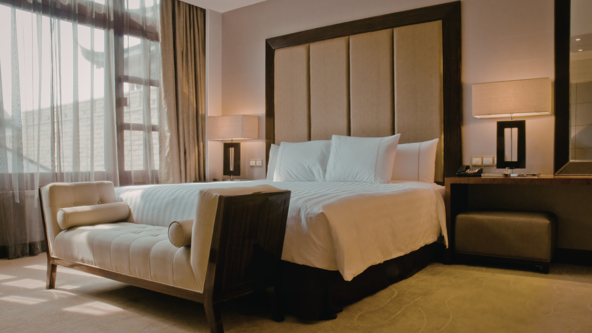 Modern hotel bedroom with a large bed, chaise lounge, bedside lamps, and floor-to-ceiling windows
