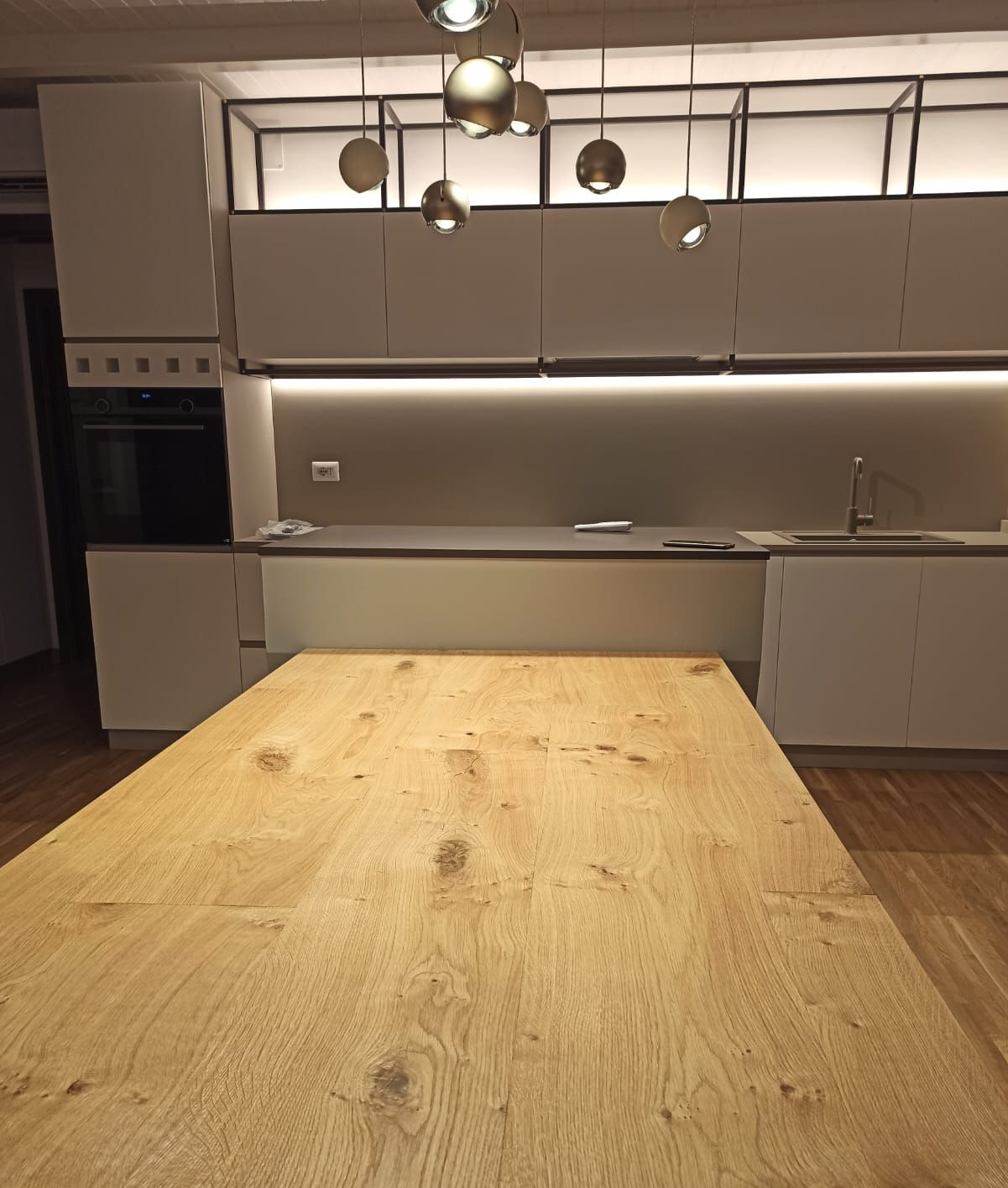 Cucina moderna con isola in legno chiaro, ripiani grigi, mobili bianchi e lampade a sospensione.