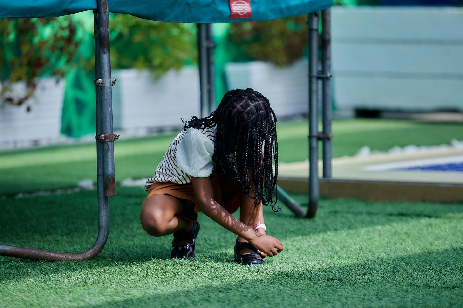 A person with braided hair crouches under a green trampoline frame on artificial turf, fastening a black sandal.