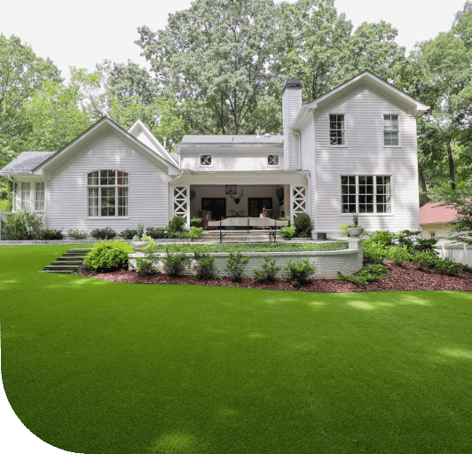 A white, two-story house with a large green lawn, a stone patio, and a covered porch, set against a backdrop of trees.