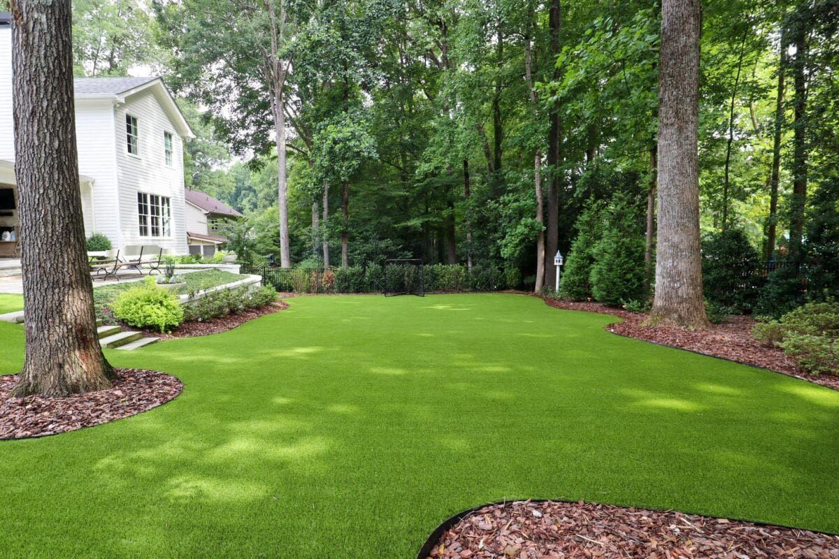 A white house with a bright green artificial turf lawn surrounded by tall trees and pine bark landscaping.
