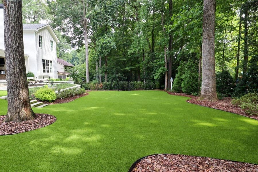 A manicured lawn of bright green artificial turf surrounded by mature trees, mulch beds, and a white house in the distance.