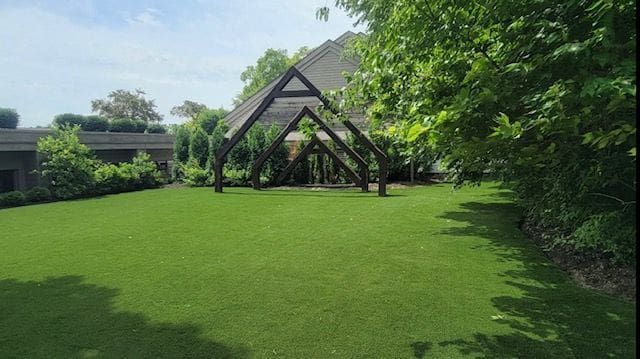 A grassy courtyard with a wooden A-frame arbor structure under a bright, sunny sky with surrounding trees.