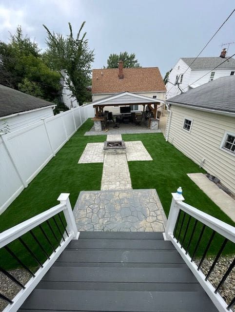 A backyard view from a deck stairs, featuring a stone path, a central fire pit, artificial turf, and a gazebo.