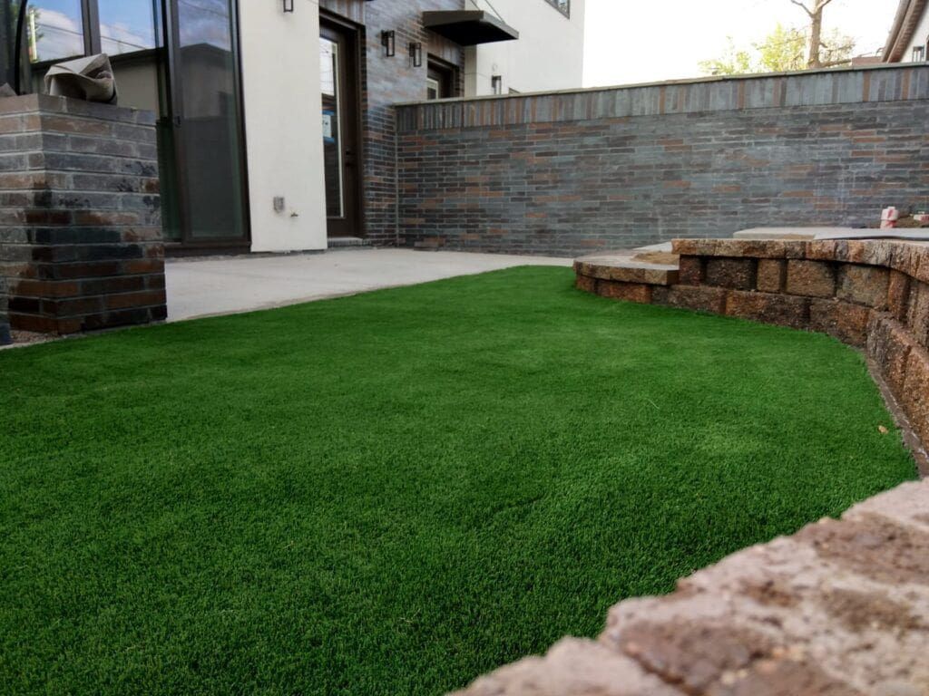 A small, manicured artificial grass lawn enclosed by stone retaining walls next to a modern patio and house exterior.