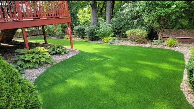 A vibrant green synthetic lawn with curved edges, bordered by gravel paths, garden plants, and a red wooden deck.