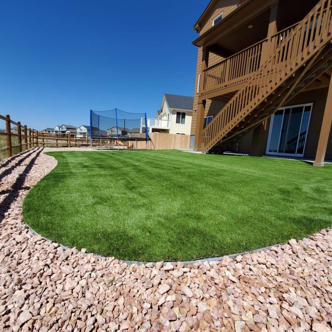 Backyard with green lawn, rock border, wooden deck stairs, and blue trampoline under a clear sky