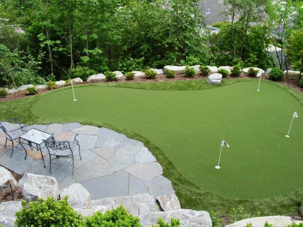 A backyard putting green with four holes next to a stone patio with a table and chairs, surrounded by lush green trees.