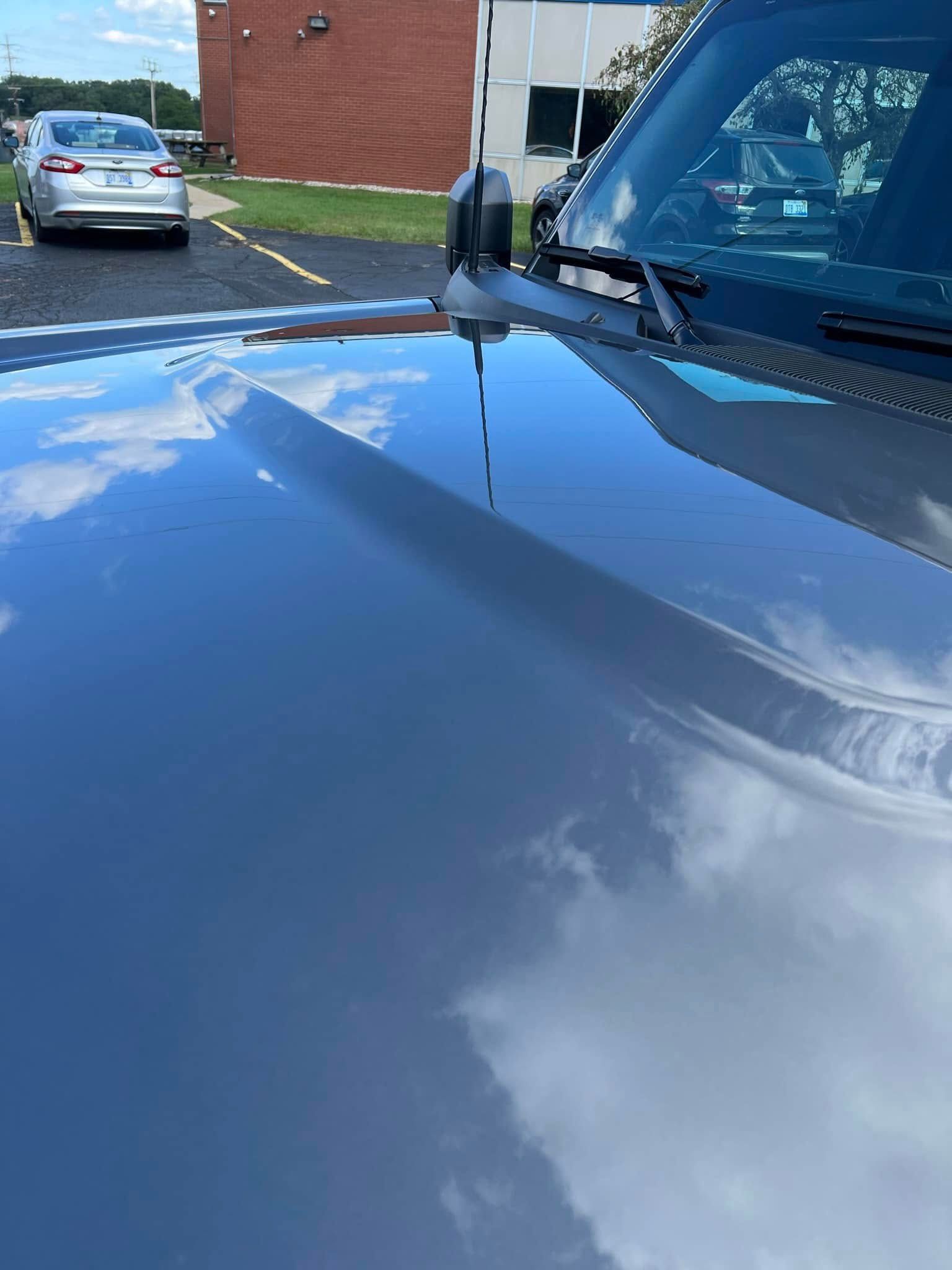 Shiny blue car hood reflecting clouds beside a brick building and parked cars