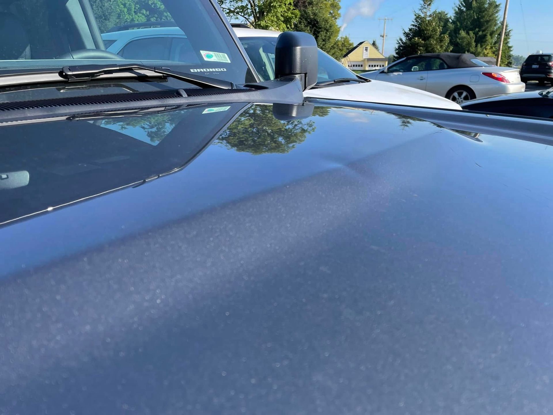Shiny blue car hood and windshield in a sunny parking lot, with trees and other cars reflected on the paint